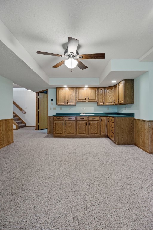 kitchen featuring brown cabinetry, wainscoting, wooden walls, light colored carpet, and a ceiling fan