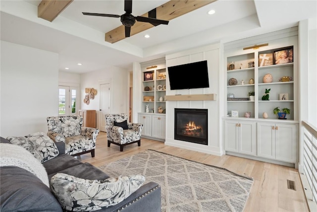 living room featuring beamed ceiling, ceiling fan, a glass covered fireplace, and light wood finished floors