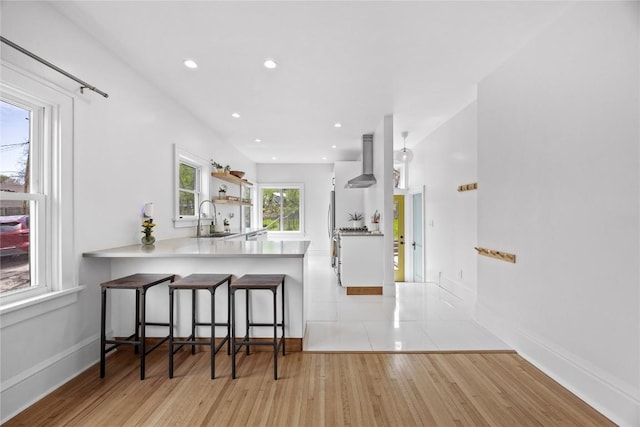 kitchen with a breakfast bar area, a sink, open shelves, light wood-type flooring, and wall chimney range hood