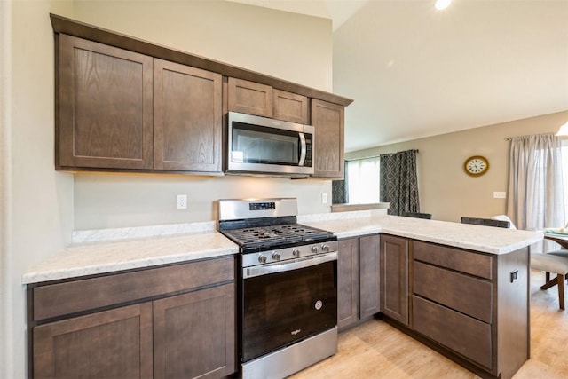 kitchen with appliances with stainless steel finishes, a peninsula, light wood-style flooring, light stone counters, and dark brown cabinets