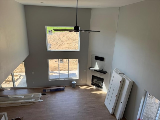 unfurnished living room featuring wood finished floors, a glass covered fireplace, a towering ceiling, and a ceiling fan