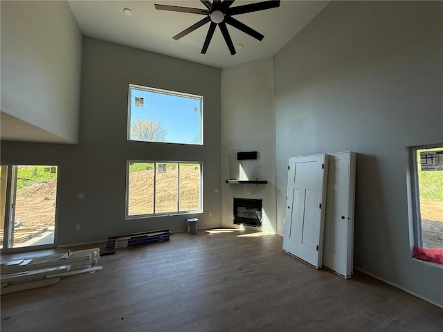 unfurnished living room with a high ceiling, a ceiling fan, wood finished floors, a glass covered fireplace, and baseboards