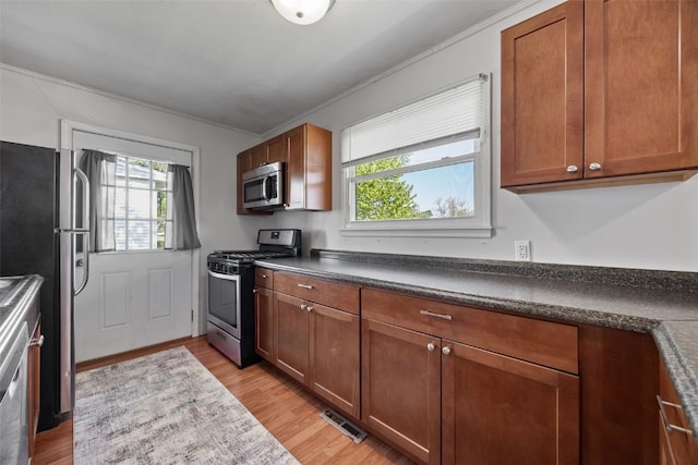 kitchen featuring appliances with stainless steel finishes, dark countertops, brown cabinets, light wood-type flooring, and crown molding