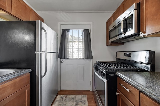 kitchen with stainless steel appliances, dark countertops, wood finished floors, brown cabinetry, and crown molding