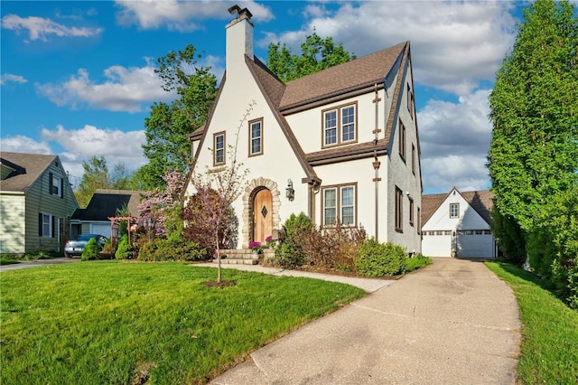 tudor home featuring stucco siding, a detached garage, a chimney, a front yard, and an outbuilding