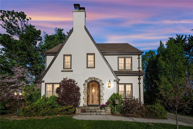 tudor home featuring stucco siding, a chimney, and roof with shingles