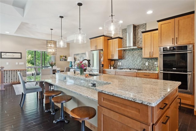 kitchen featuring wall chimney range hood, appliances with stainless steel finishes, dark wood-style floors, tasteful backsplash, and a large island with sink