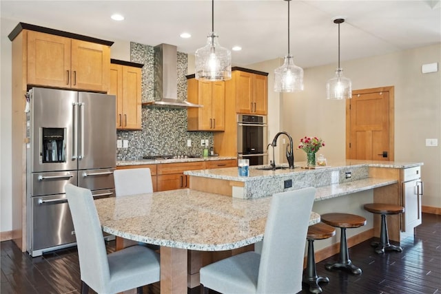 kitchen with stainless steel appliances, wall chimney exhaust hood, dark wood-style flooring, a sink, and a center island with sink