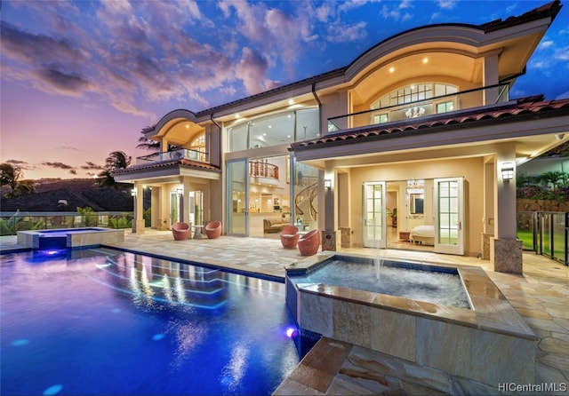 back house at dusk featuring a balcony, a swimming pool with hot tub, pool water feature, and a patio