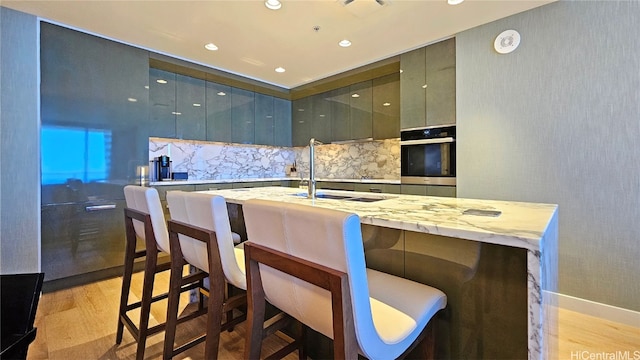 kitchen featuring oven, light hardwood / wood-style floors, light stone counters, decorative backsplash, and a breakfast bar area