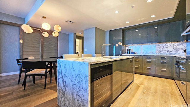 kitchen featuring backsplash, hanging light fixtures, wine cooler, light hardwood / wood-style flooring, and a kitchen island with sink