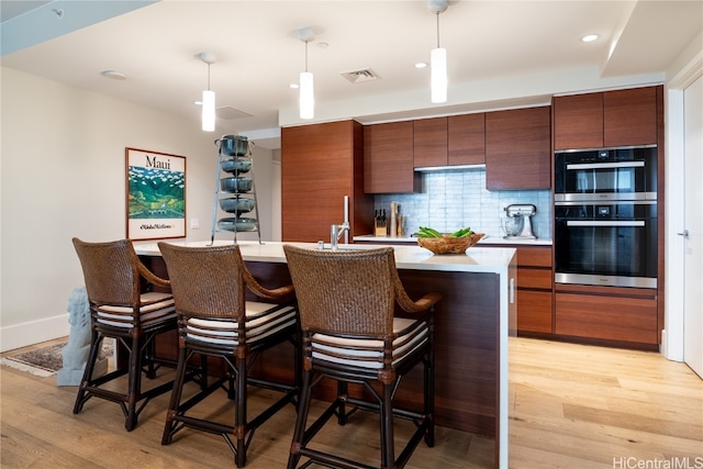 kitchen featuring tasteful backsplash, double oven, hanging light fixtures, light hardwood / wood-style floors, and a kitchen island with sink