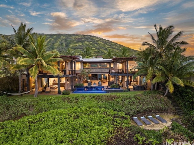 back house at dusk with a balcony, a mountain view, a patio area, and a pool with hot tub