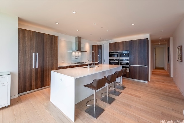 kitchen featuring wall chimney range hood, sink, light wood-type flooring, a breakfast bar, and a center island with sink