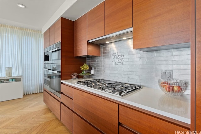 kitchen featuring brown cabinets, under cabinet range hood, backsplash, appliances with stainless steel finishes, and light countertops