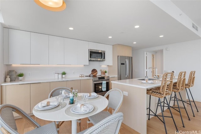 kitchen featuring an island with sink, appliances with stainless steel finishes, tasteful backsplash, light hardwood / wood-style floors, and white cabinetry