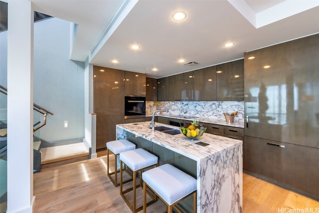 kitchen featuring oven, a kitchen breakfast bar, an island with sink, sink, and light wood-type flooring