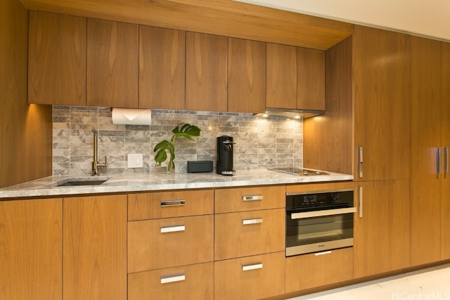 kitchen with oven, backsplash, sink, cooktop, and light stone counters