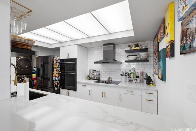 kitchen featuring wall chimney range hood, black appliances, light stone countertops, white cabinets, and tasteful backsplash