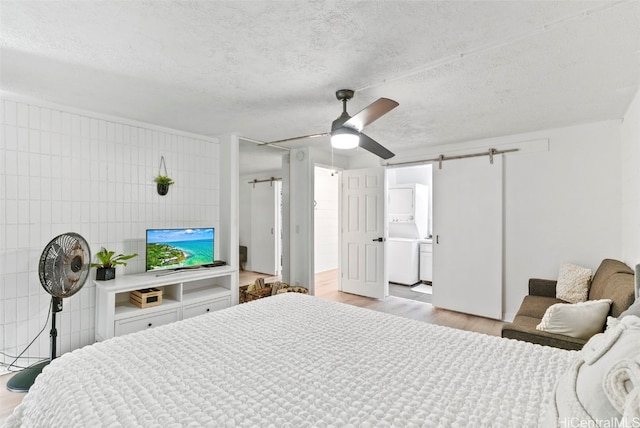 bedroom featuring ensuite bathroom, a barn door, ceiling fan, stacked washer and dryer, and light hardwood / wood-style flooring