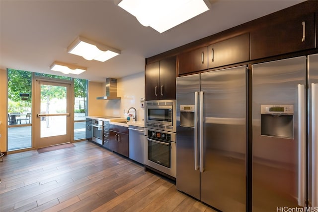 kitchen with sink, light wood-type flooring, dark brown cabinets, wall chimney exhaust hood, and built in appliances