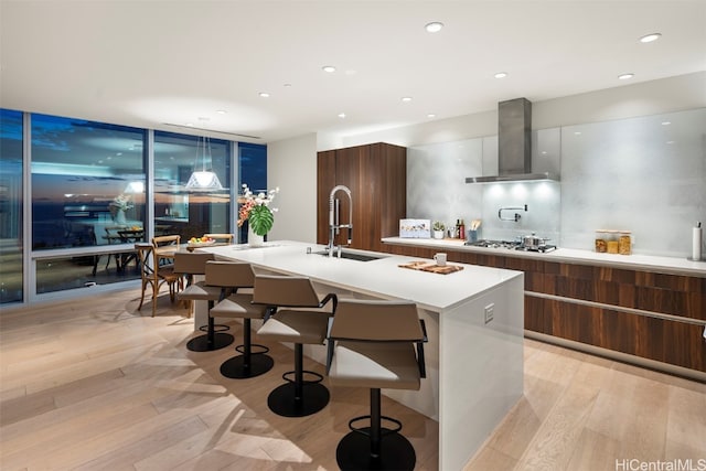 kitchen with a breakfast bar area, a kitchen island with sink, light hardwood / wood-style floors, wall chimney exhaust hood, and sink