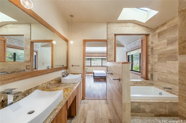 bathroom with a relaxing tiled tub, hardwood / wood-style floors, tile walls, vanity, and lofted ceiling with skylight