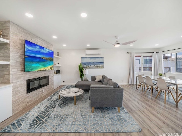 living room featuring a wall mounted AC, a stone fireplace, light hardwood / wood-style floors, and ceiling fan