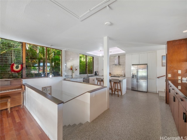 kitchen featuring a kitchen bar, appliances with stainless steel finishes, decorative backsplash, wall chimney exhaust hood, and white cabinetry