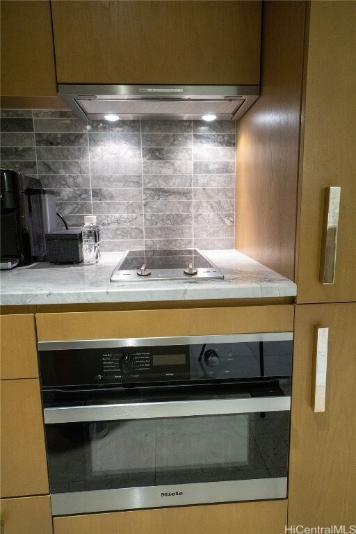 interior details with range hood, light stone counters, decorative backsplash, and oven