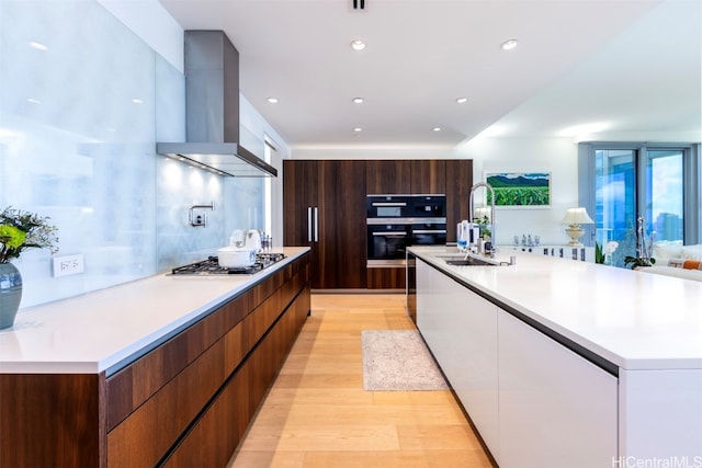 kitchen with wall chimney range hood, double oven, a large island, stainless steel gas stovetop, and light hardwood / wood-style flooring