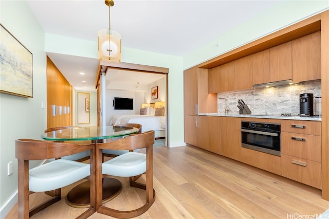 kitchen with oven, backsplash, hanging light fixtures, range hood, and light hardwood / wood-style flooring