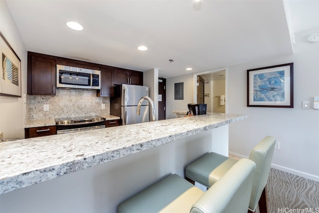 kitchen featuring dark brown cabinets, stainless steel appliances, backsplash, light stone countertops, and a kitchen bar