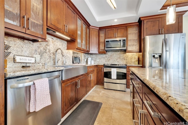 kitchen featuring appliances with stainless steel finishes, a tray ceiling, decorative light fixtures, light stone counters, and decorative backsplash