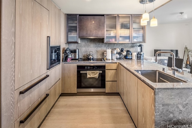 kitchen with light stone counters, stainless steel oven, light brown cabinetry, sink, and decorative light fixtures