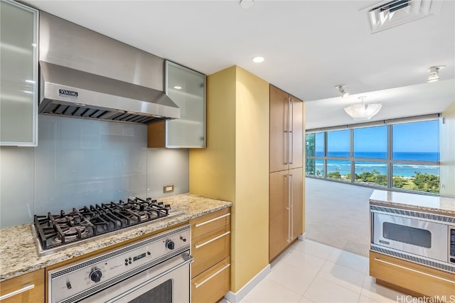 kitchen featuring light stone counters, light tile patterned floors, expansive windows, wall chimney exhaust hood, and stainless steel appliances