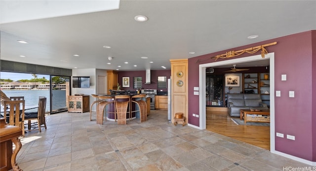 kitchen with a water view, wall chimney exhaust hood, stainless steel stove, ceiling fan, and a breakfast bar