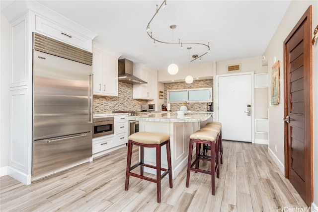 kitchen featuring white cabinets, appliances with stainless steel finishes, light hardwood / wood-style flooring, pendant lighting, and wall chimney exhaust hood