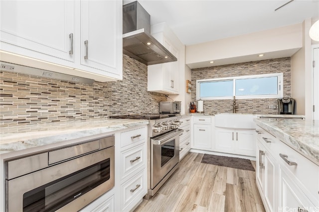 kitchen with light stone countertops, appliances with stainless steel finishes, light wood-type flooring, wall chimney exhaust hood, and white cabinets