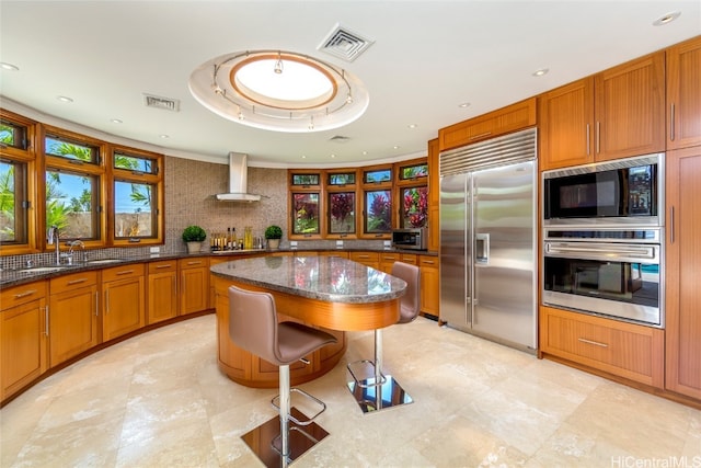 kitchen featuring a kitchen island, dark stone countertops, a breakfast bar area, built in appliances, and wall chimney exhaust hood