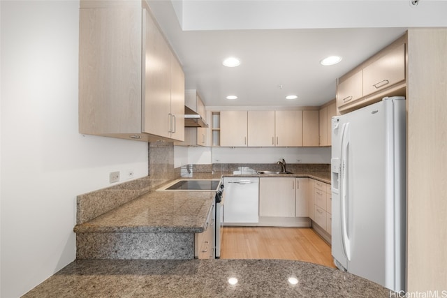 kitchen featuring white appliances, wall chimney range hood, sink, light wood-type flooring, and dark stone countertops