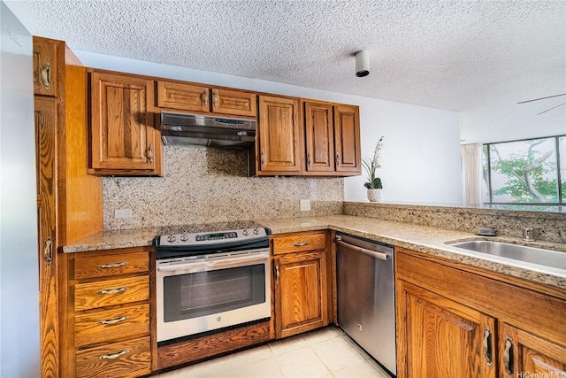 kitchen with under cabinet range hood, brown cabinetry, stainless steel appliances, and a textured ceiling