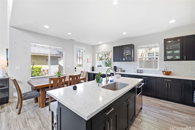 kitchen featuring decorative backsplash, an island with sink, sink, light stone countertops, and light wood-type flooring