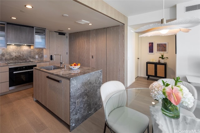 kitchen featuring oven, sink, backsplash, light hardwood / wood-style floors, and a kitchen island with sink