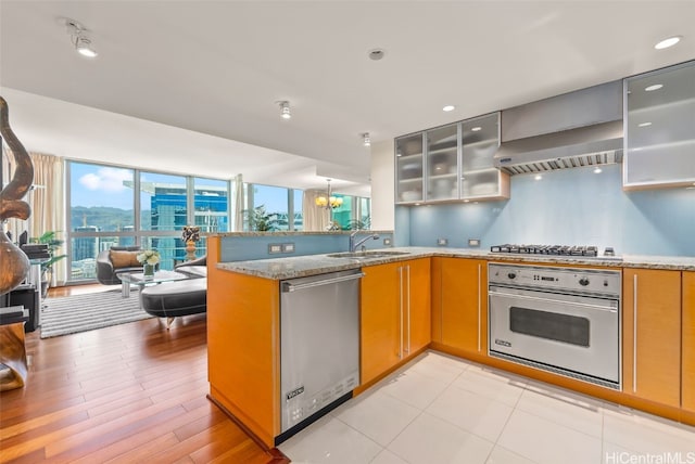 kitchen with wall chimney range hood, light stone countertops, kitchen peninsula, and appliances with stainless steel finishes