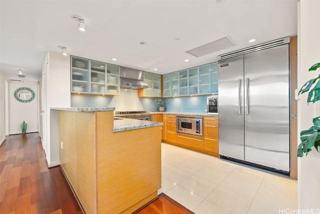 kitchen with wall chimney exhaust hood, light stone counters, built in appliances, kitchen peninsula, and backsplash