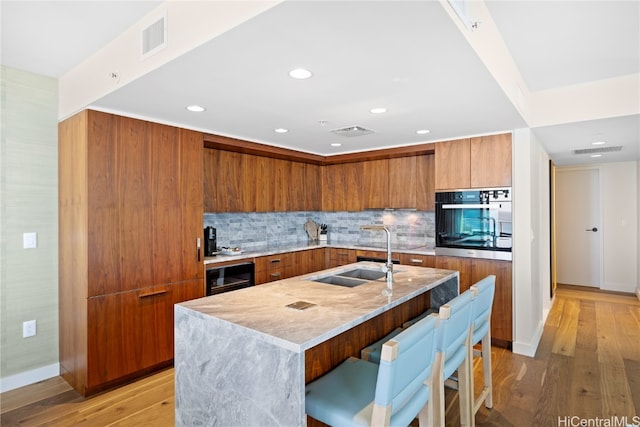 kitchen with decorative backsplash, a kitchen island with sink, oven, sink, and light wood-type flooring