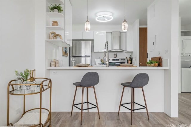 kitchen featuring wall chimney exhaust hood, stainless steel appliances, stacked washer / drying machine, decorative light fixtures, and white cabinets