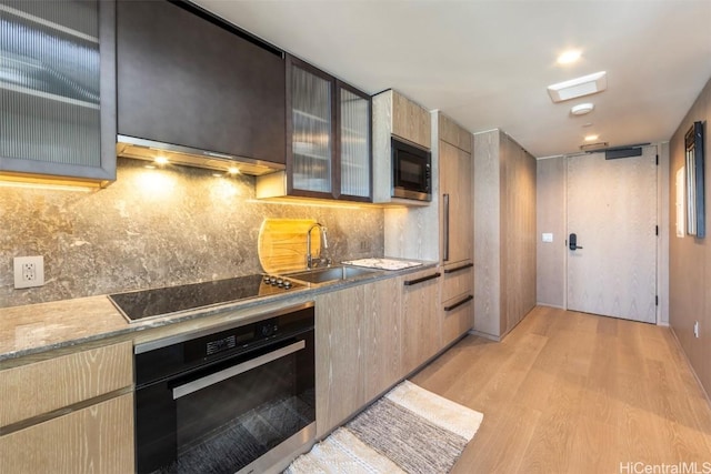 kitchen featuring light wood-style flooring, under cabinet range hood, a sink, decorative backsplash, and black appliances