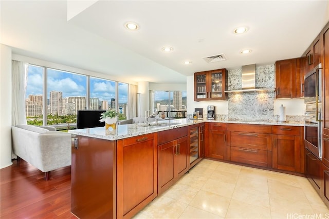 kitchen with wall chimney exhaust hood, tasteful backsplash, wine cooler, light stone counters, and kitchen peninsula